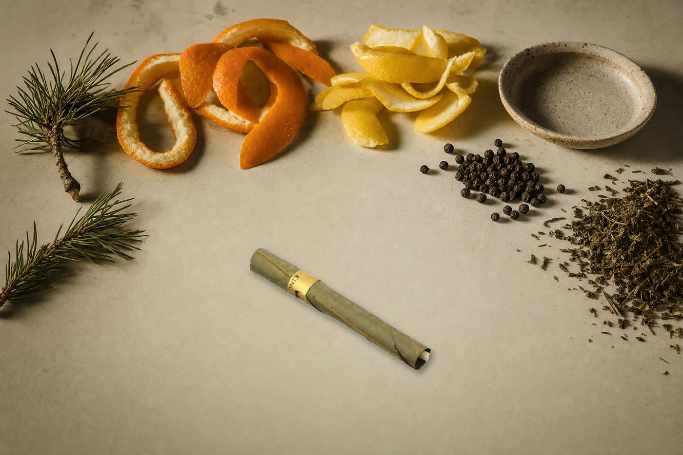Citrus peel, pine sprigs and peppercorns arranged on a cream surface beside unlit King Palm rolls